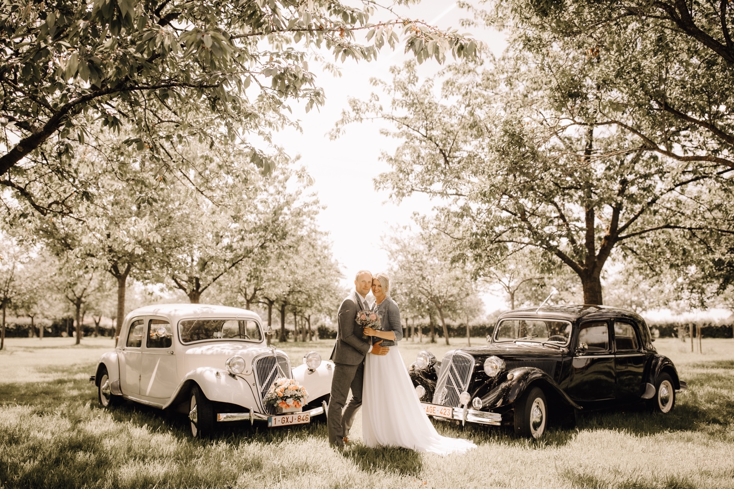 Bruidspaar poseert tijdens fotoshoot naast twee oldtimers in een mooie boomgaard