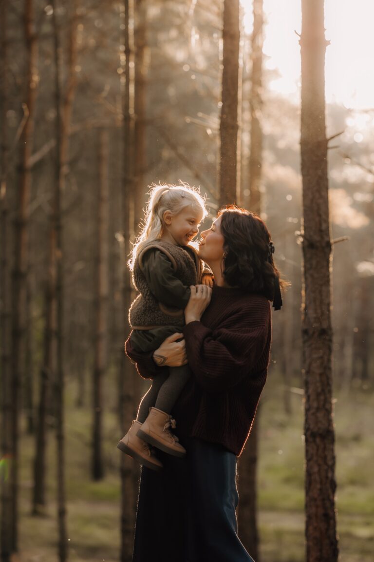 Motherhood sessie in het bos - moeder en dochter kijken elkaar liefdevol aan tussen de bomen