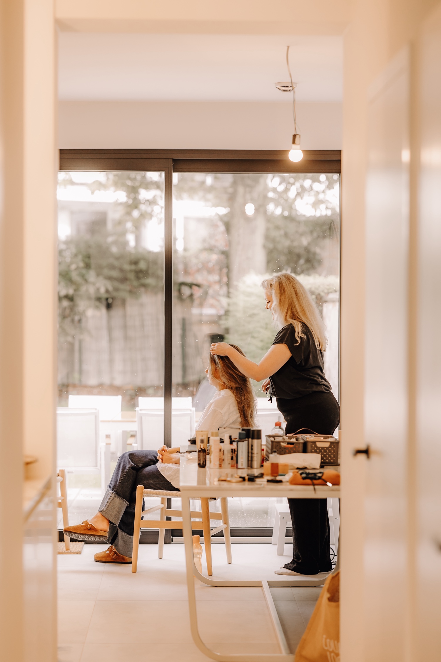 Een regenachtige huwelijksdag bij Hoeve Engelendael - bruid zit voor een groot raam en laat haar make-up en haar doen