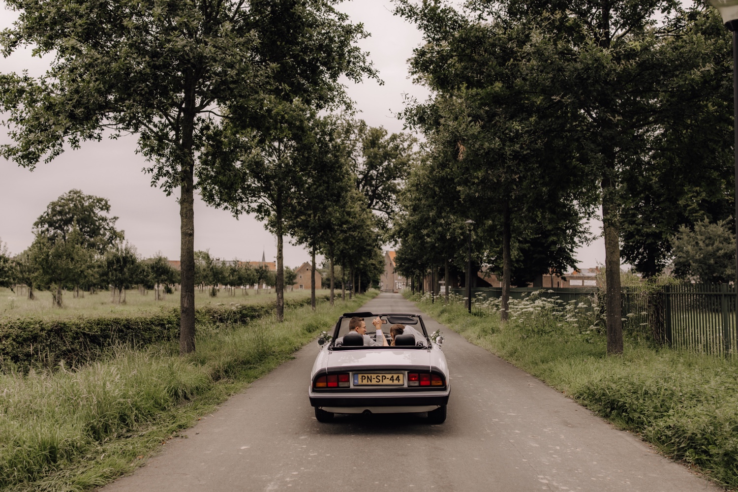 Intiem huwelijk bij La Butte Aux Bois - bruidspaar rijdt door bomenlaan met hun witte oldtimer